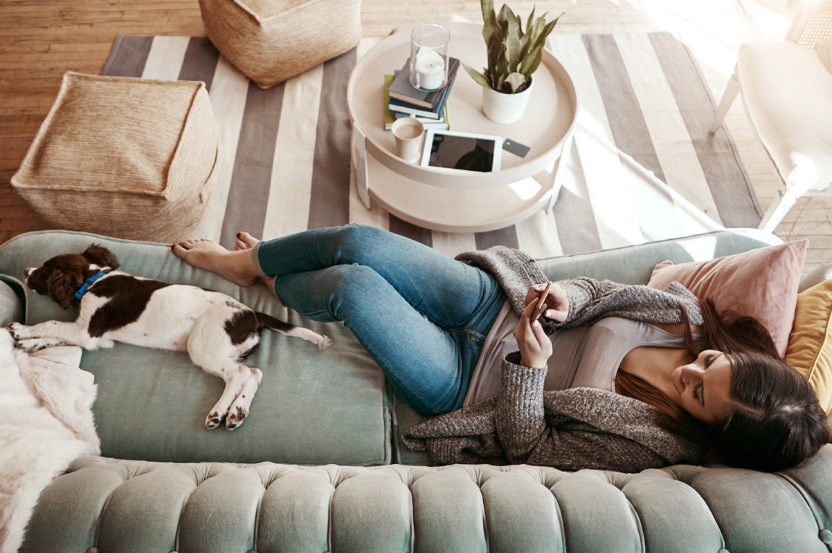 Living room with wood floor, coffee table and woman laying on couch with dog, texting on phone