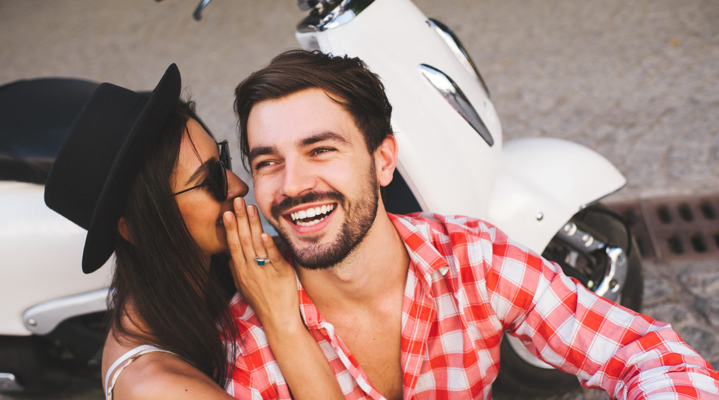 Woman whispering to laughing man with parked scooter behind them
