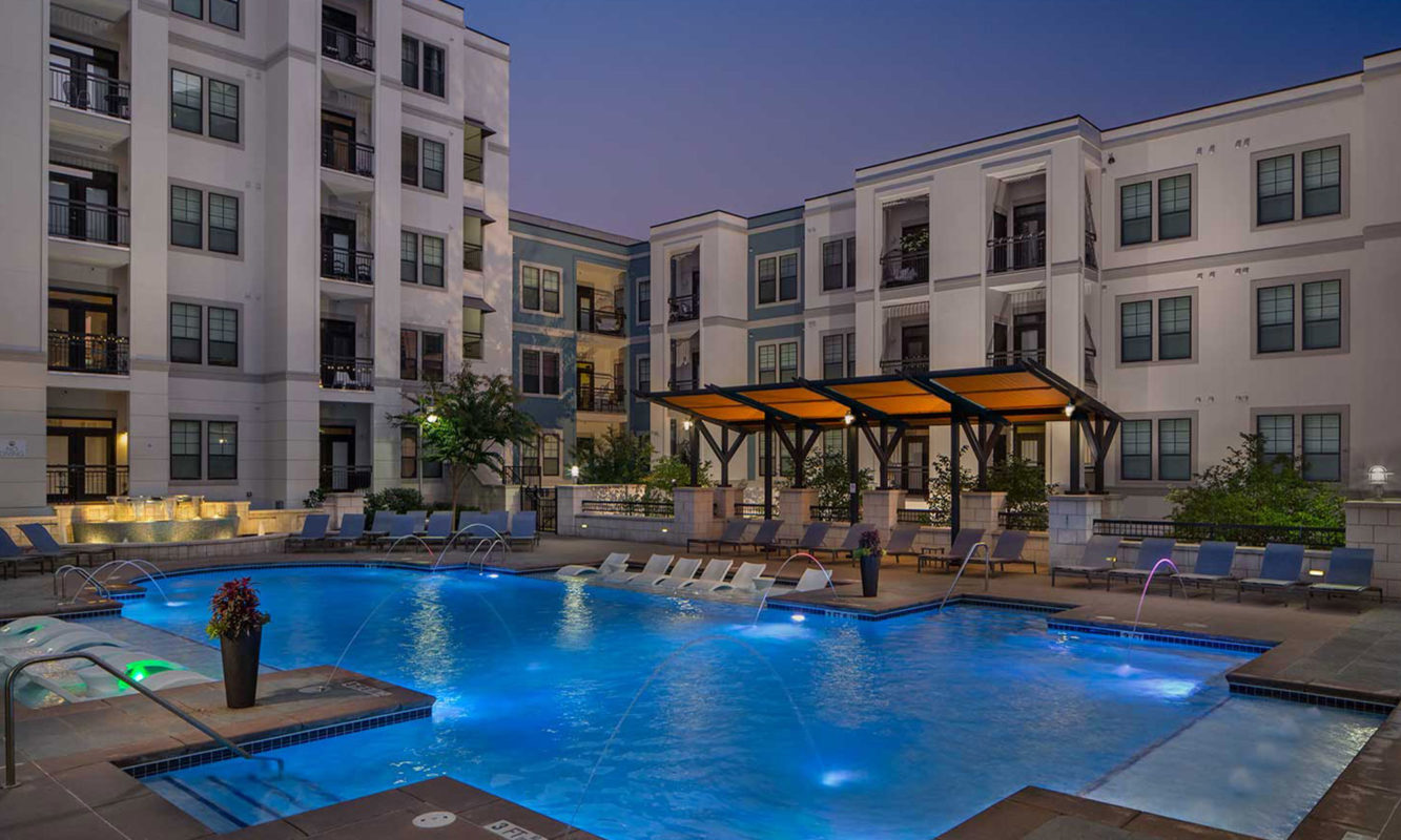 Dusk shot of pool with in-water lounge seats, additional lounge chairs, lighted fountain and covered area