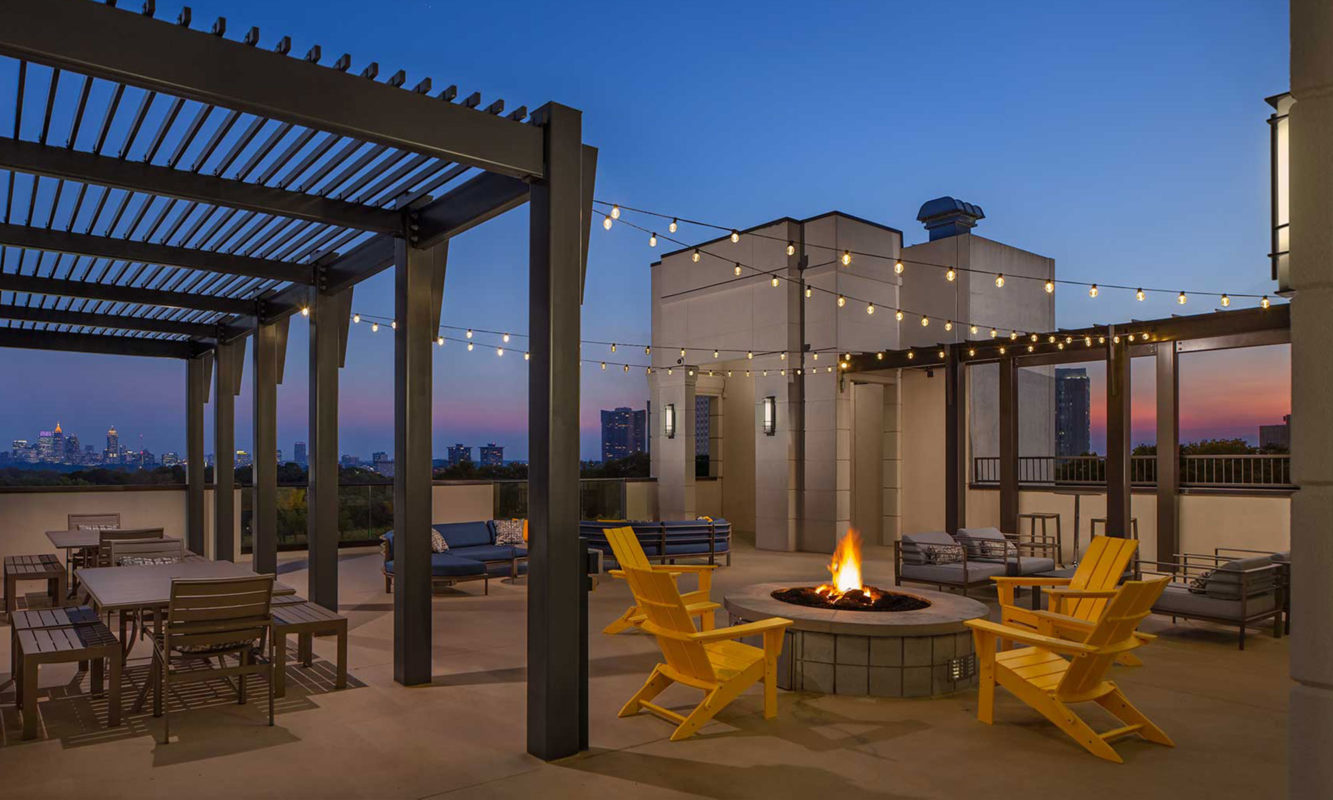 Dusk shot of rooftop fire pit with string lighting and tables and chairs